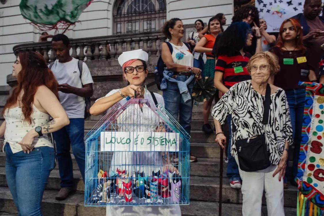 A woman, dressed as a nurse, holds a bird cage containing a sign that says 'Louco è o Sistema'